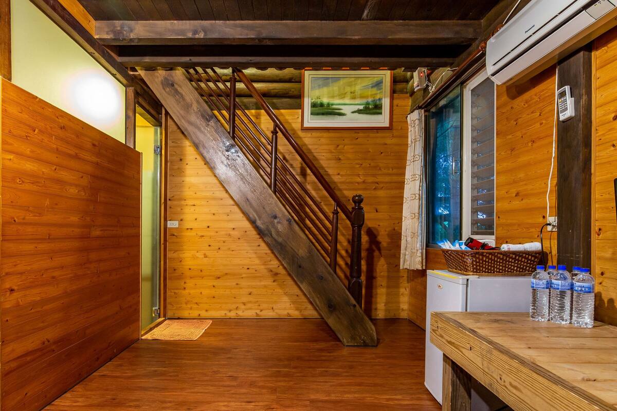 Ein Holzinnenraum mit Treppe, einem gerahmten Bild, einem Fenster mit Jalousien und einem kleinen Kühlschrank mit Wasserflaschen.