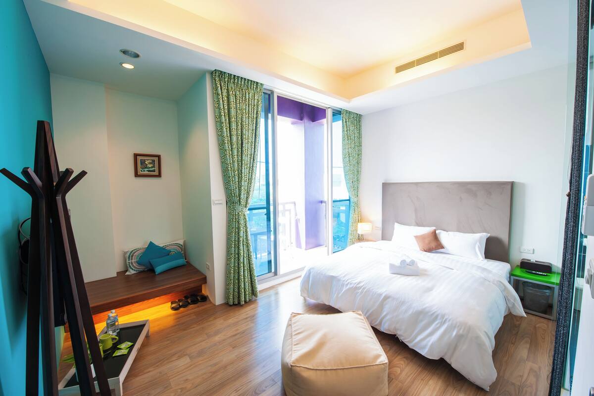Une chambre d’hôtel moderne dotée d’un grand lit, d’un coin salon avec un banc et des coussins, d’une petite table avec un plateau, et d’un balcon avec des rideaux.
