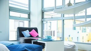 Ein modernes Wohnzimmer mit Ledersofa, blau-rosa Kissen, einem Glastisch und Blick auf Gebäude durch große Fenster.