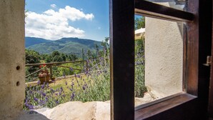 Uitzicht door een raam op een landelijk gebouw met een stenen muur, een houten balk en een berglandschap met lavendelvelden.