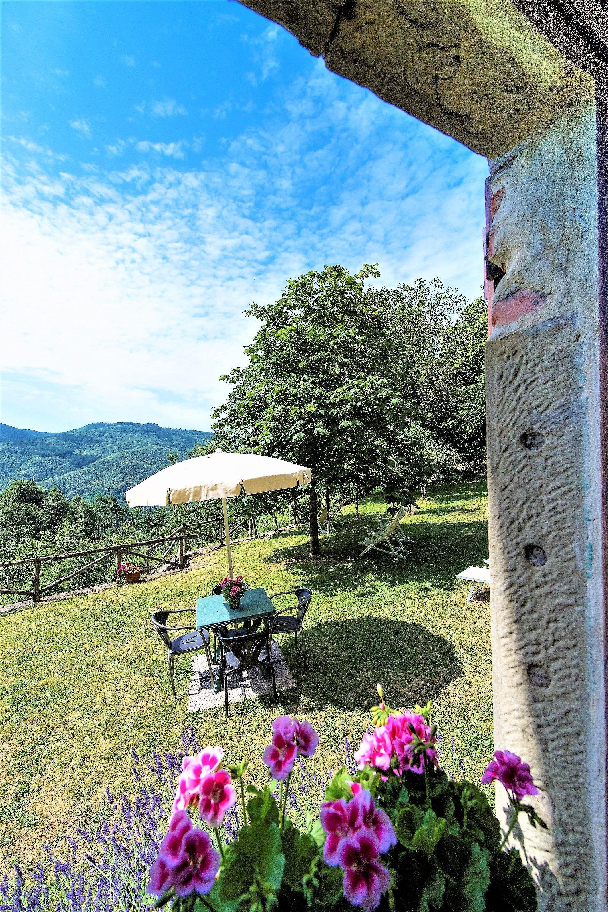 Une terrasse avec une table et des chaises, un parasol blanc et une vue sur des collines verdoyantes.