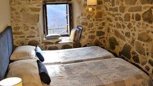Een slaapkamer met stenen muren, een raam, een bed, kussens, een bedlamp en een boekenplank.