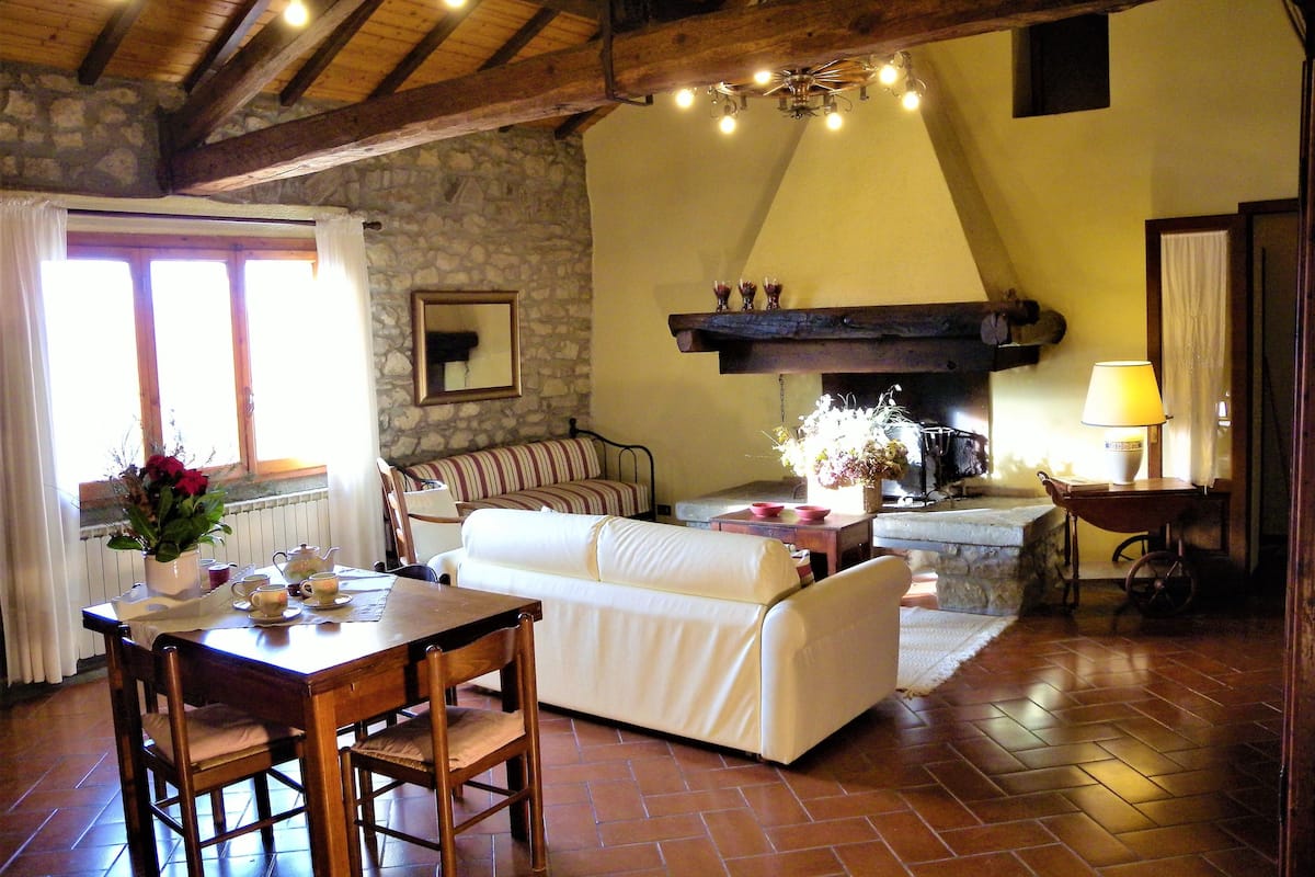 Una sala de estar acogedora con pared de piedra, vigas de madera, chimenea, comedor con mesa y sillas, y un sofá.