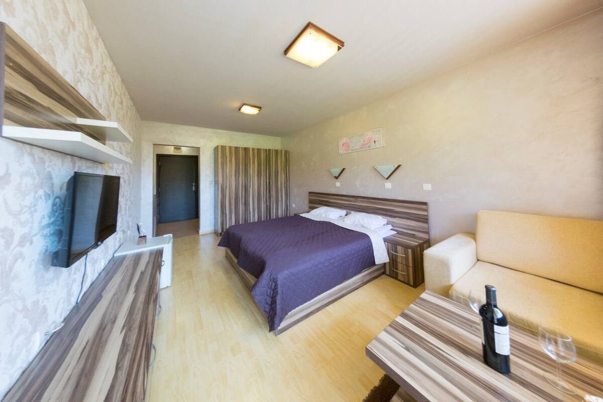 A hotel room with a bed, bedside table with a bottle and glass, a TV mounted on the wall, and a sofa.