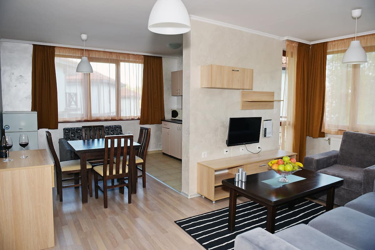 A modern living room with a flat-screen TV, a dining area with wooden furniture, and a kitchen with a microwave.
