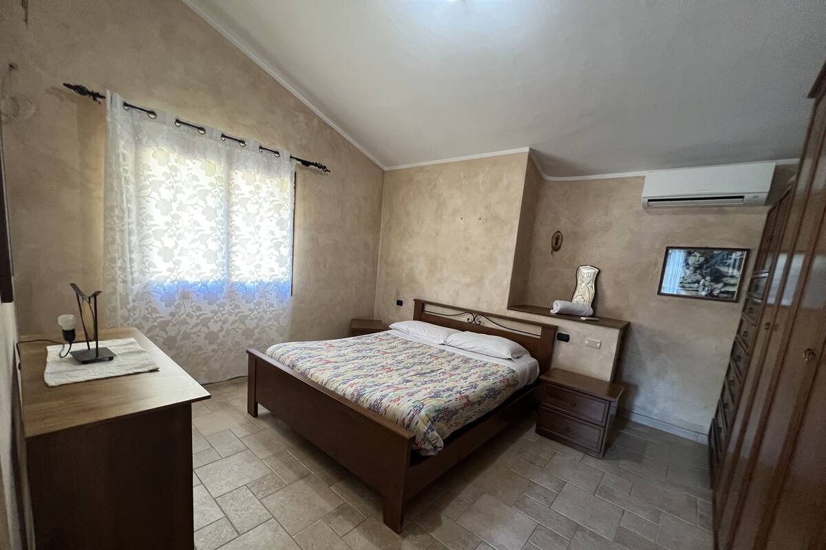 Una camera da letto con un letto, comodini, una scrivania, una finestra con tende, un condizionatore d'aria e un quadro appeso al muro.