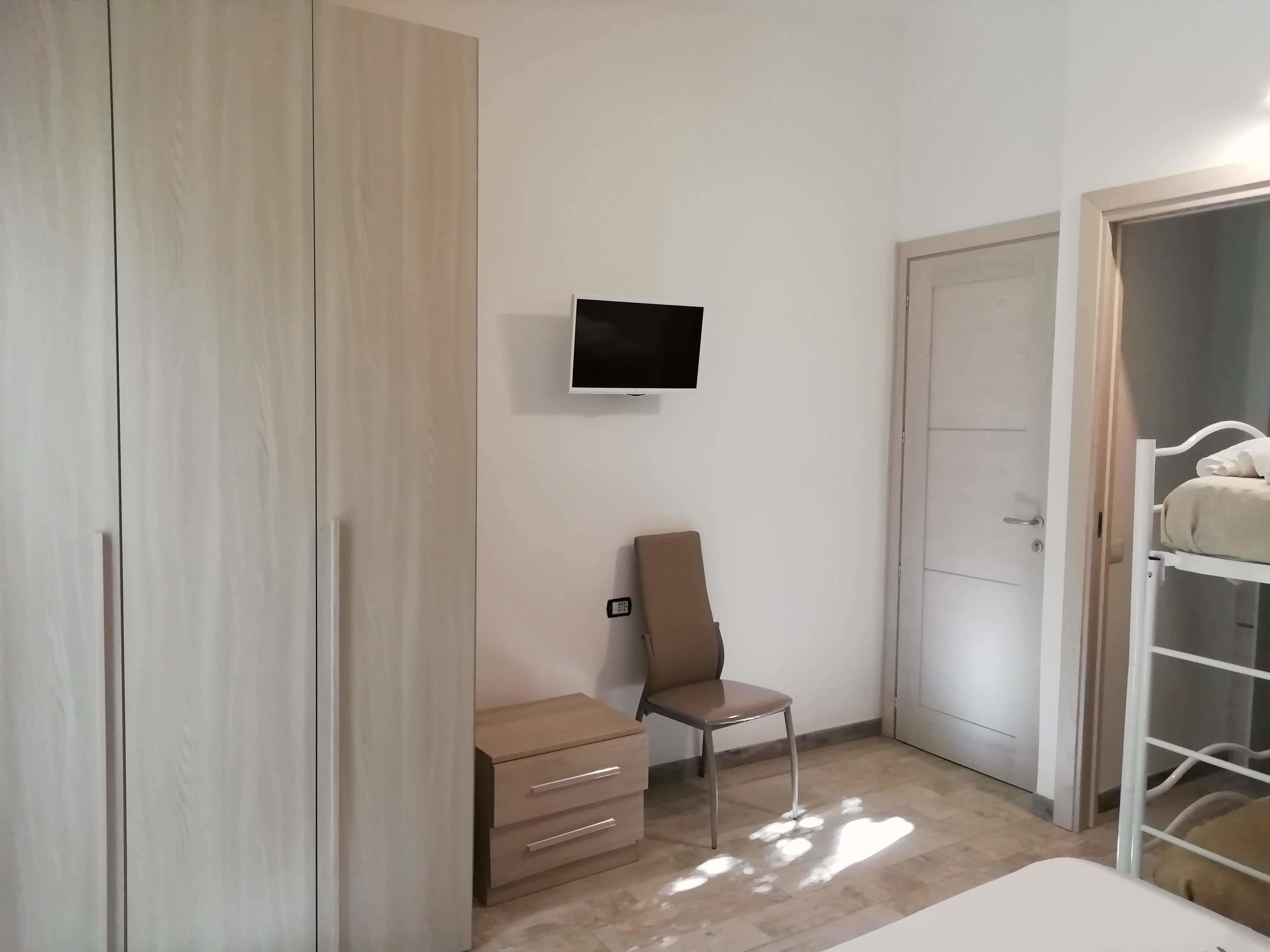 A bedroom with a bed, a TV mounted on the wall, a chair, a wardrobe, and a door.