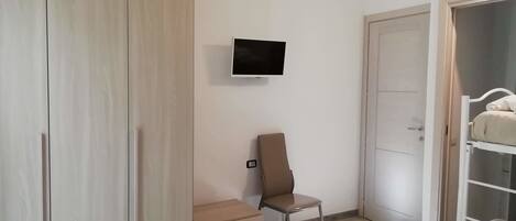 A bedroom with a bed, a TV mounted on the wall, a chair, a wardrobe, and a door.