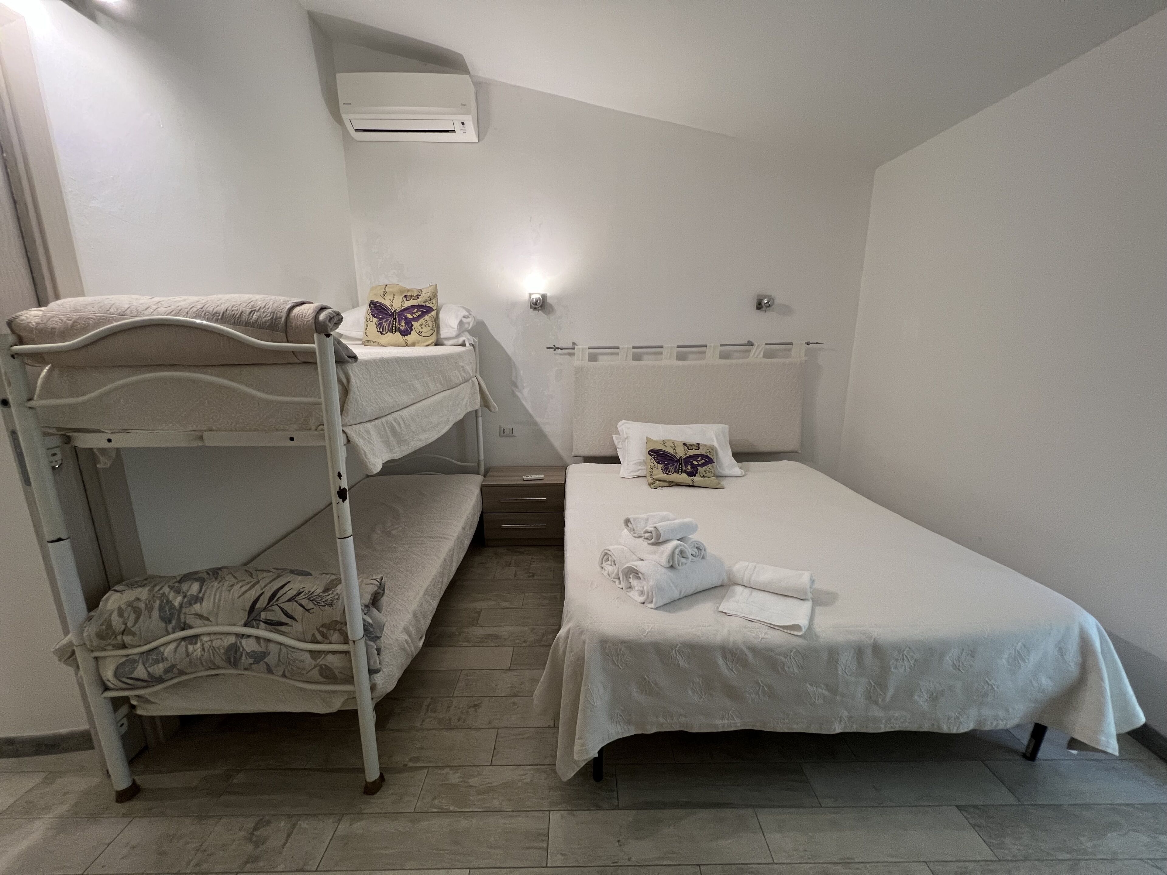 A single-bed room with a bunk bed on the left, a bedside table with a lamp, and a wall-mounted air conditioner.