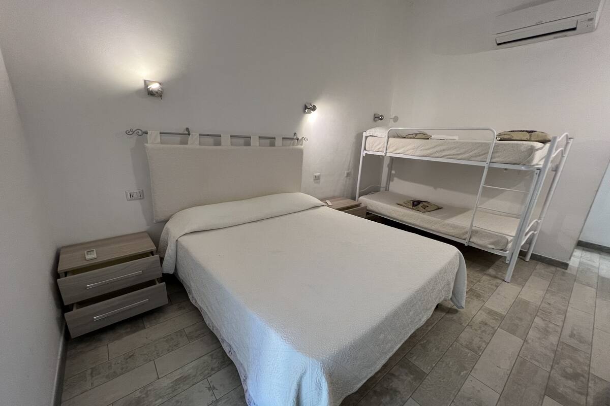Un letto singolo con testiera, un comodino con cassetti, un piccolo comodino con una lampada e un letto a castello con due piani.
