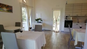 A dining area with a table set for two, a kitchen with modern appliances, and a view of the outdoors through large windows. - In Campagna da Nino (Olbia)