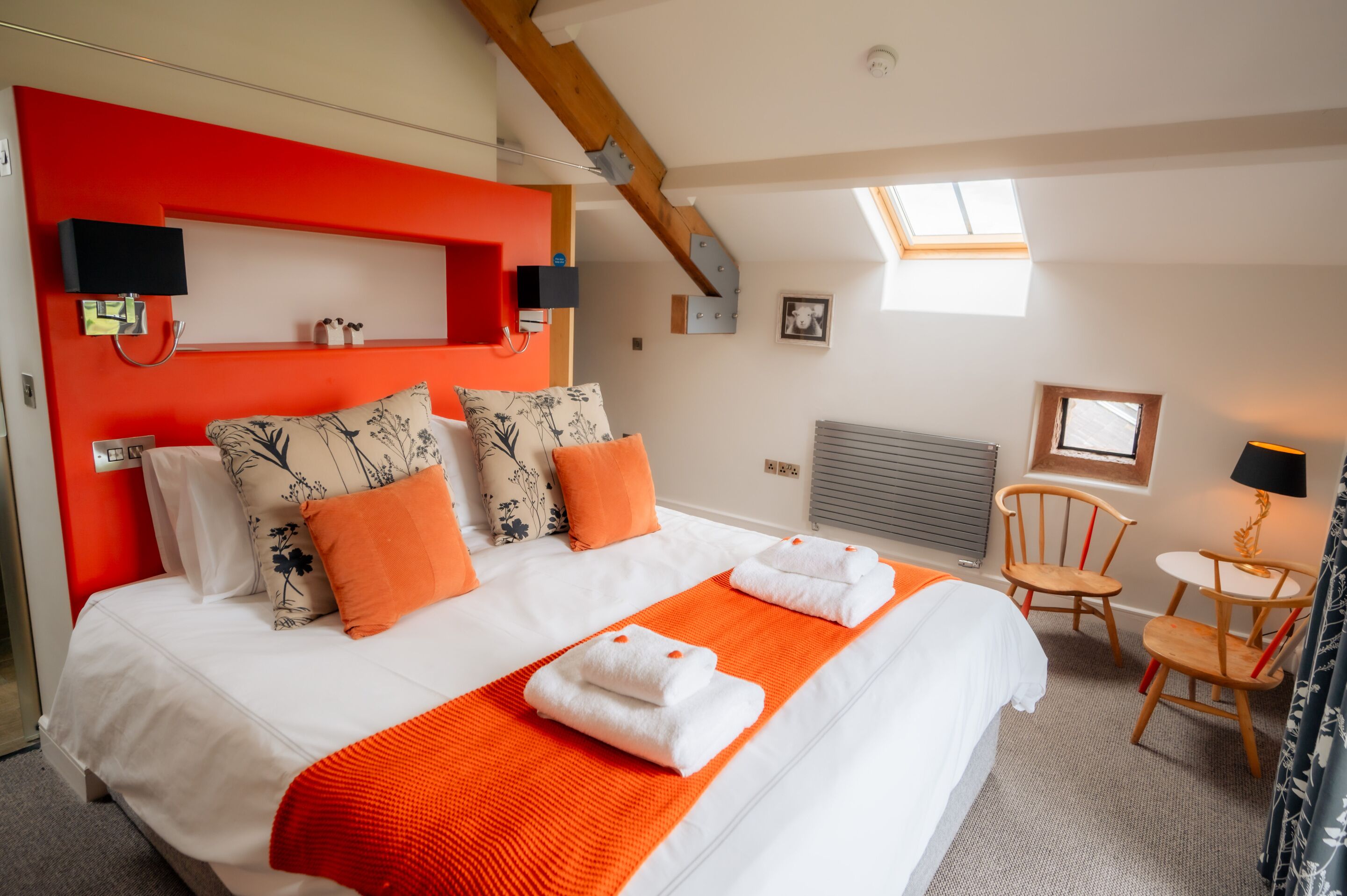 Une chambre d’hôtel avec un grand lit, des touches de couleur orange, une lucarne, une petite table avec des chaises et une lampe de chevet.