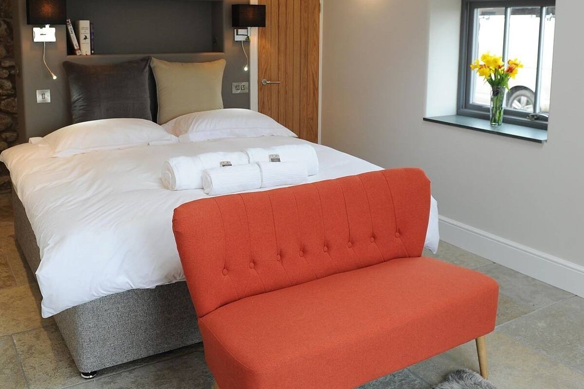 A neatly made bed with white linens and rolled towels, a red upholstered chair, and a window with a vase of flowers.