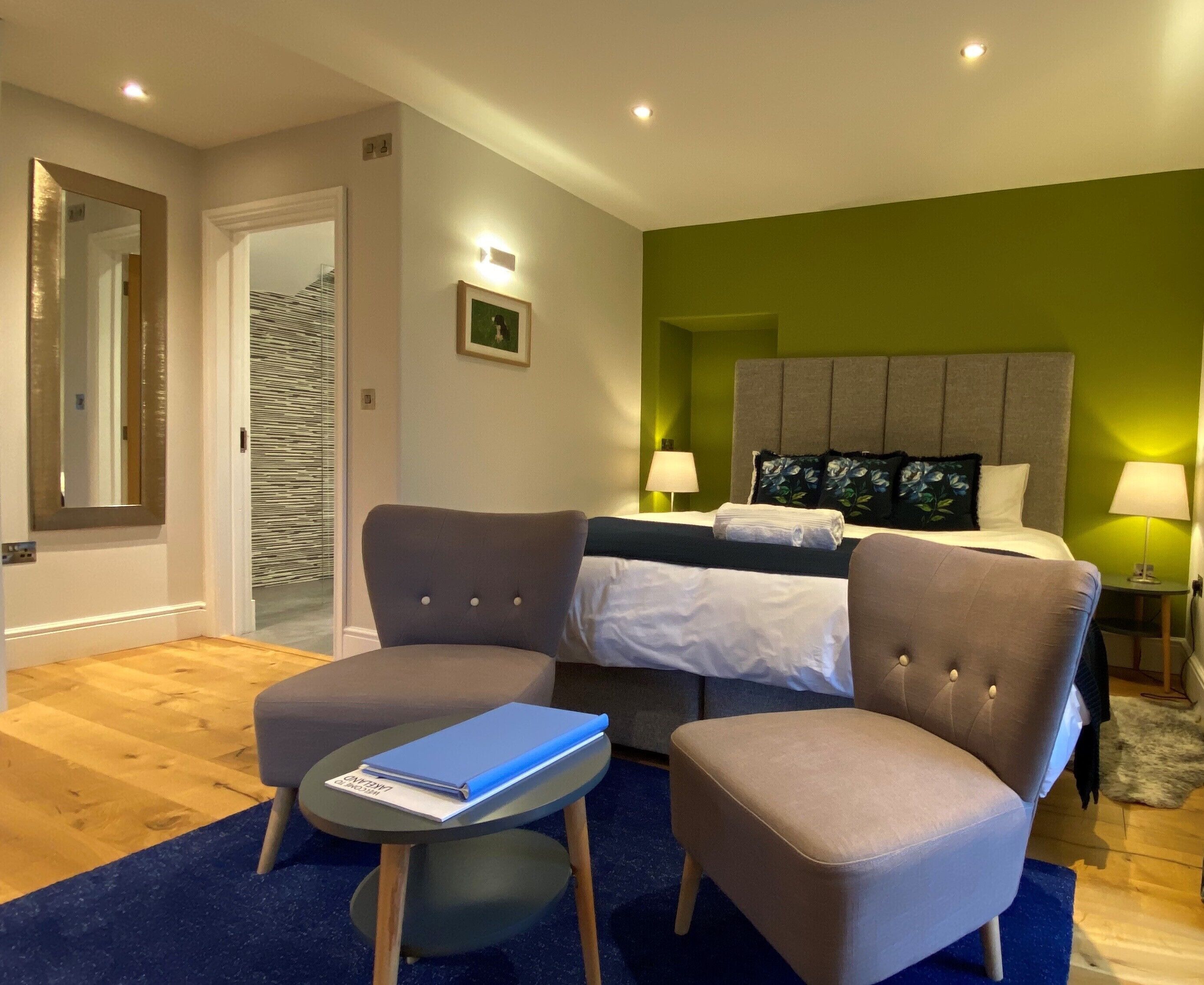 A hotel room with a bed, two armchairs, a small table with a book, a mirror, and a green accent wall.