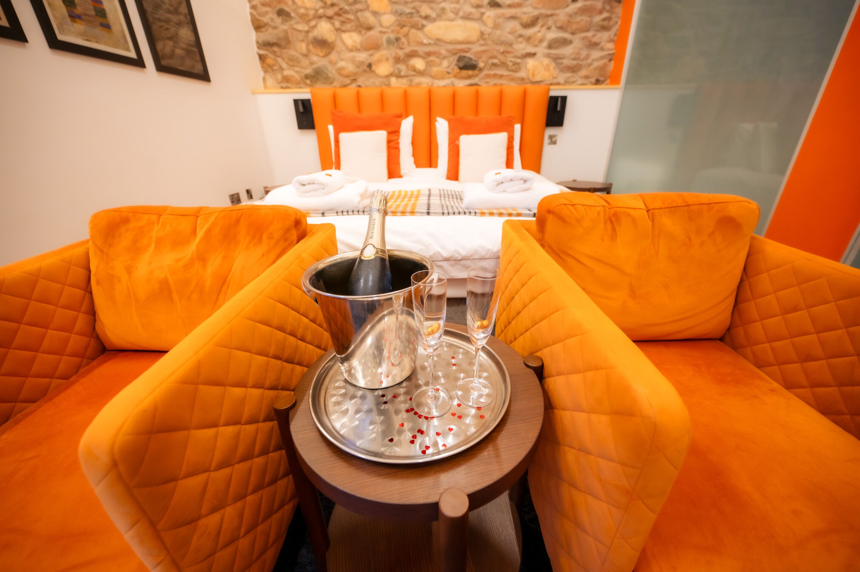 Una habitación con una cama, sillones tapizados en naranja, una bandeja con una botella de champán y copas, y una pared de piedra.