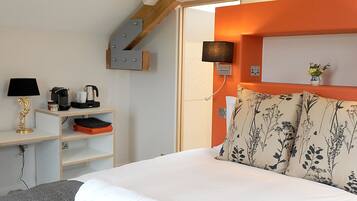 A neatly made bed with two rolled towels, a bedside table with a lamp, a coffee maker, and a shelf with folded clothes.