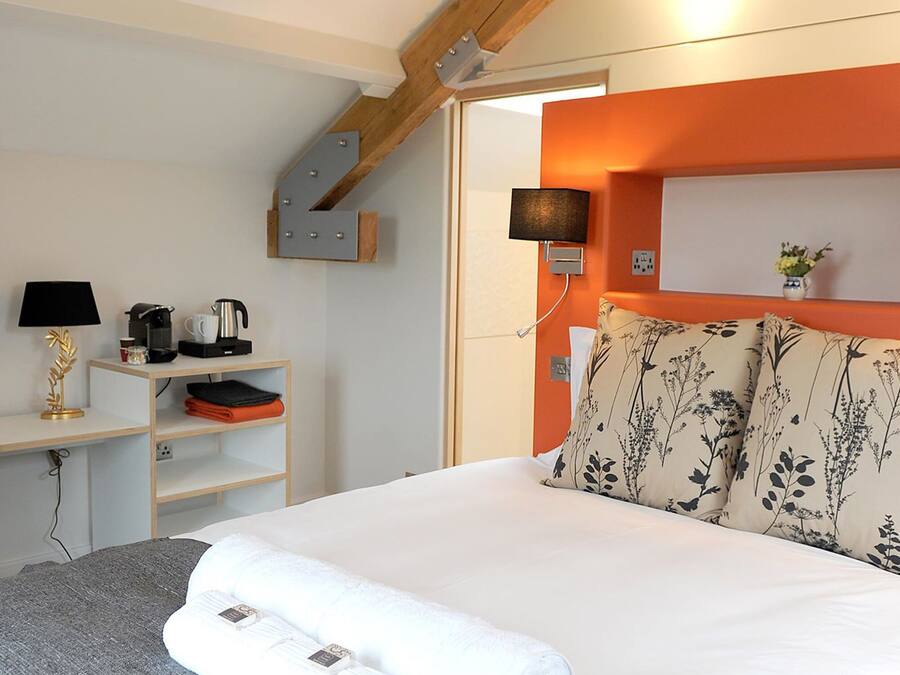 A neatly made bed with two rolled towels, a bedside table with a lamp, a coffee maker, and a shelf with folded clothes.