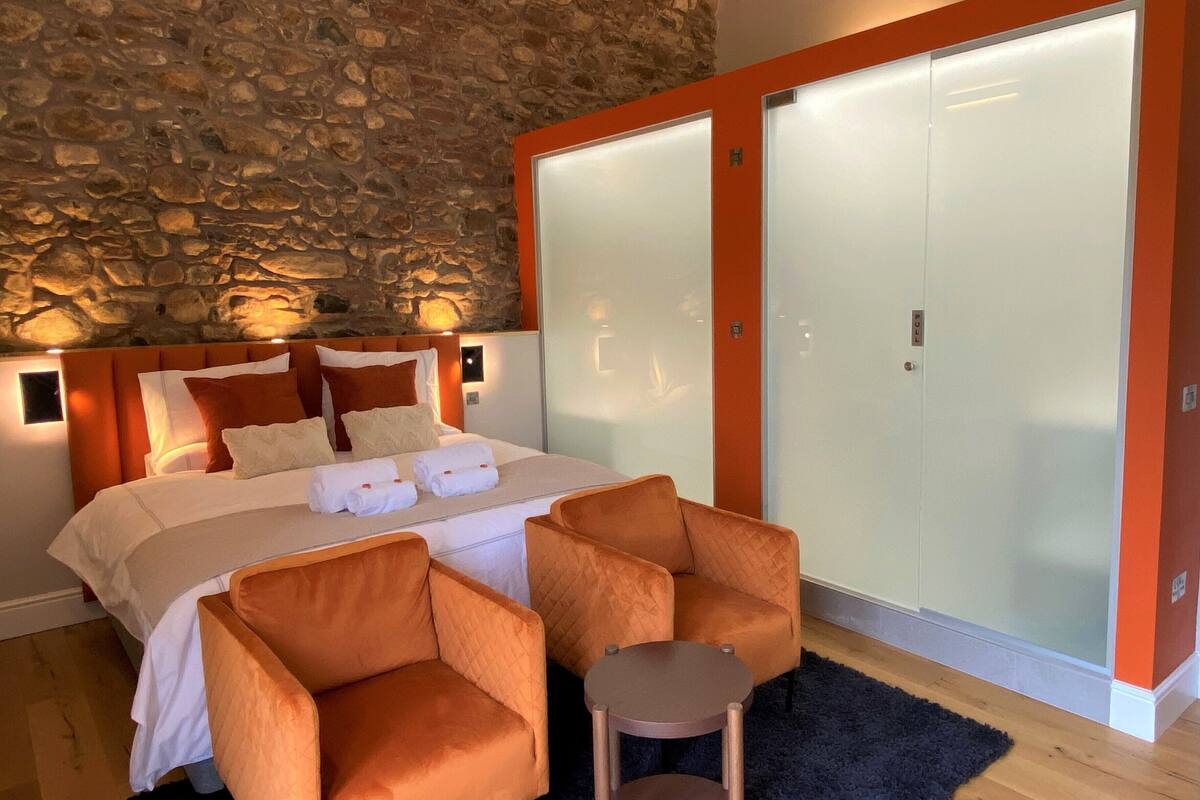 A bedroom with a stone wall, a bed with white linens, orange chairs, and a small table.