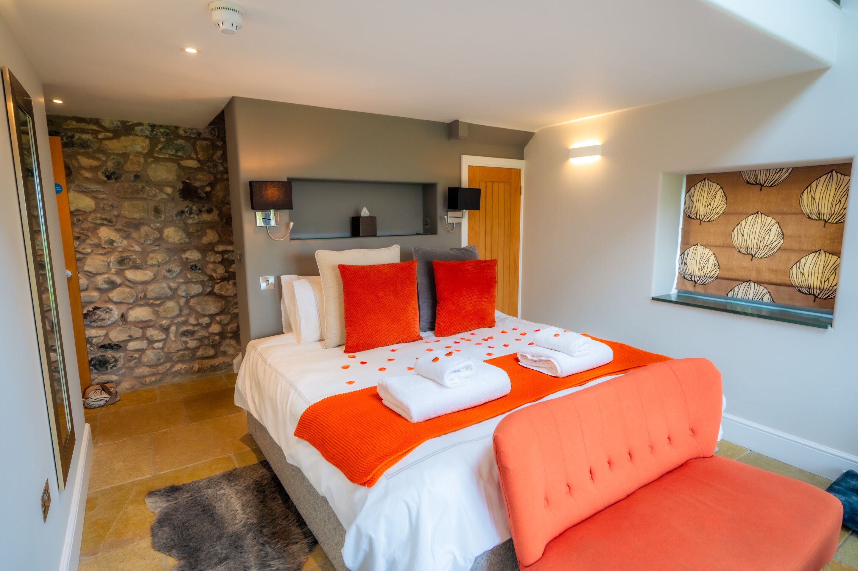 Une chambre avec un mur en pierre, un lit avec une literie blanche et des touches de couleur orange, une chaise rouge et une œuvre d’art accrochée au mur.