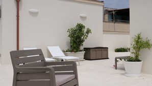 Un patio en la azotea con una silla reclinable gris, una maceta blanca con una planta y una pared blanca con una ventana y una lámpara blanca.