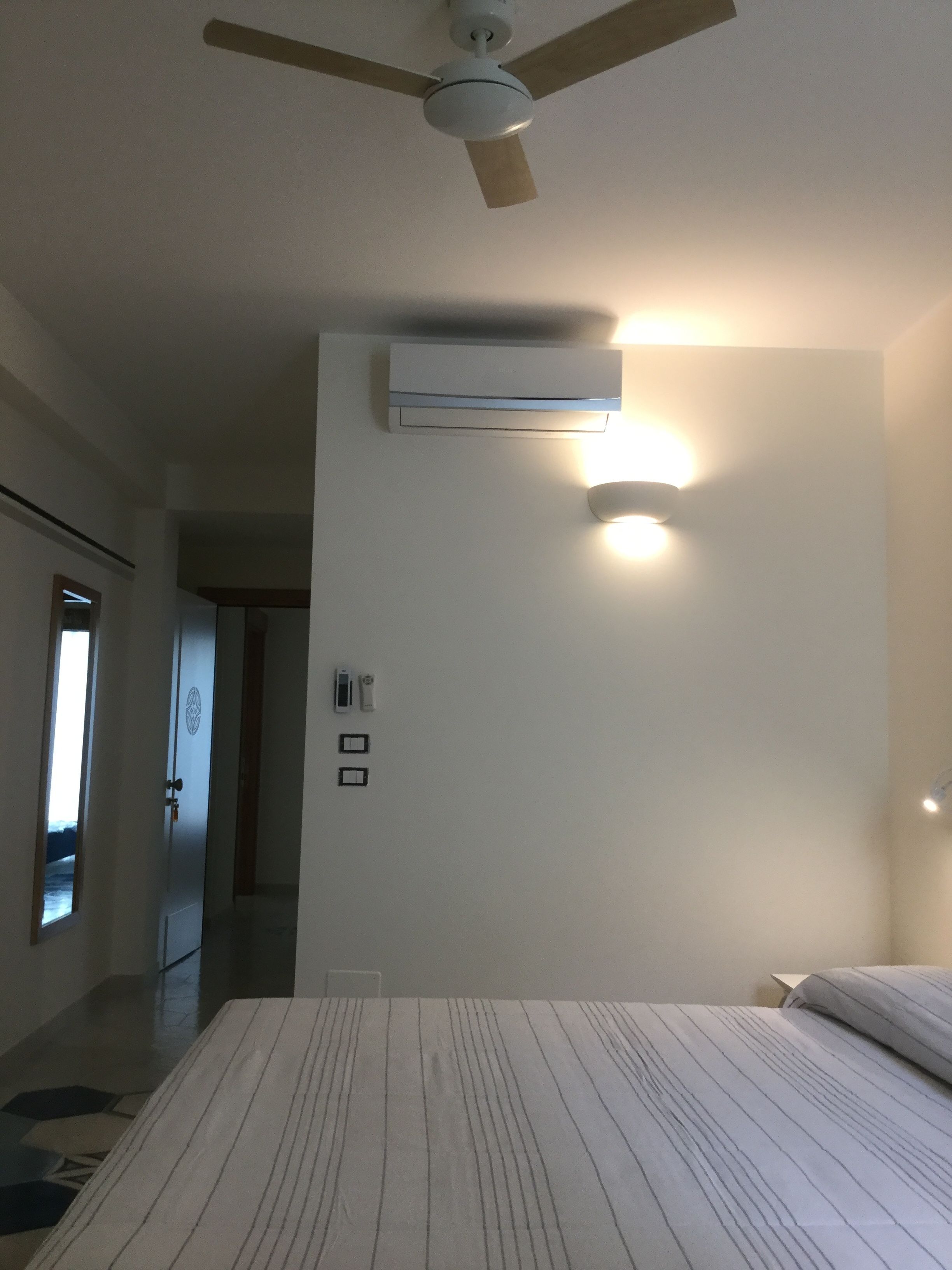 A bedroom with a bed, a ceiling fan, an air conditioning unit, a wall-mounted light, and a door with a handle.