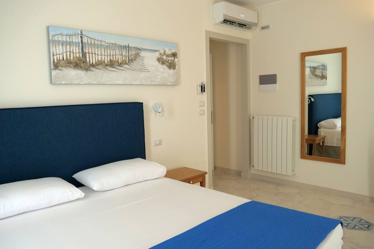 A hotel room with a large bed, a painting on the wall, a mirror, and a radiator.
