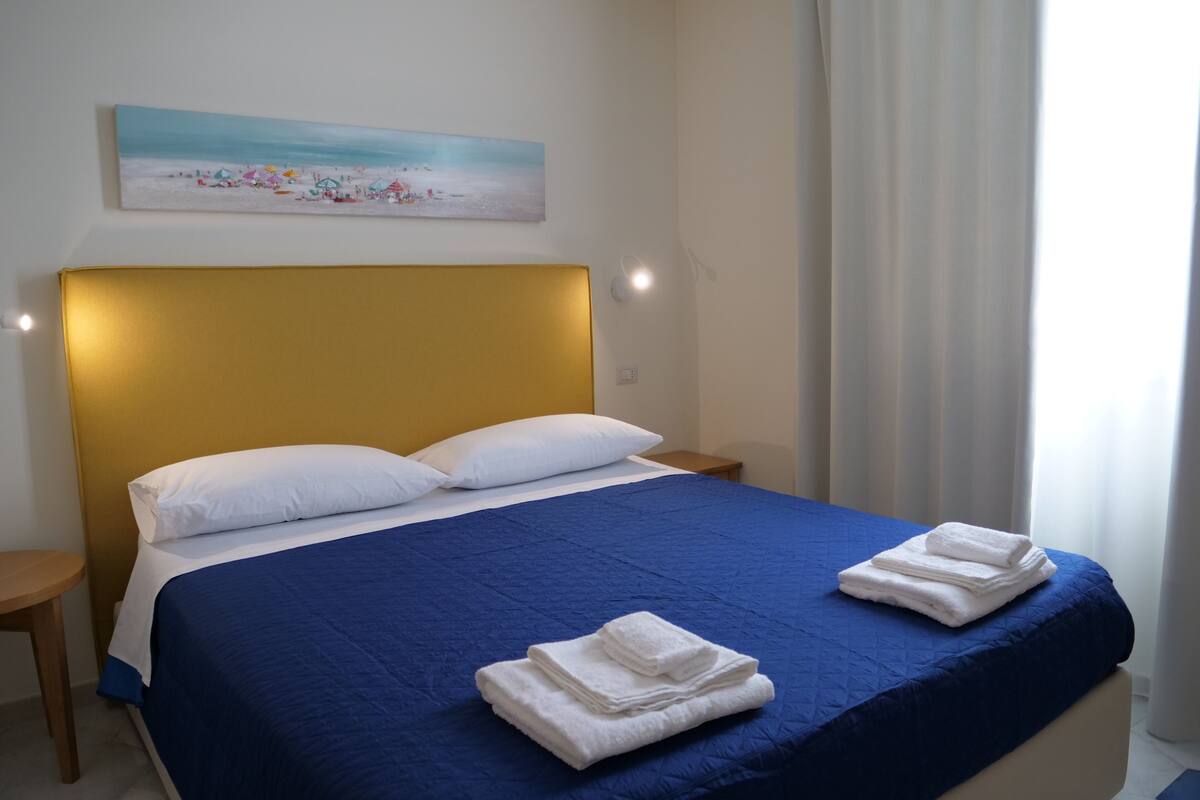 A neatly made bed with a blue bedspread and white pillows, a small wooden nightstand, and a painting of a beach scene on the wall.
