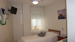 Un dormitorio con dos camas, un ventilador de techo, un televisor montado en la pared, una ventana con cortinas translúcidas y un cuadro en la pared.