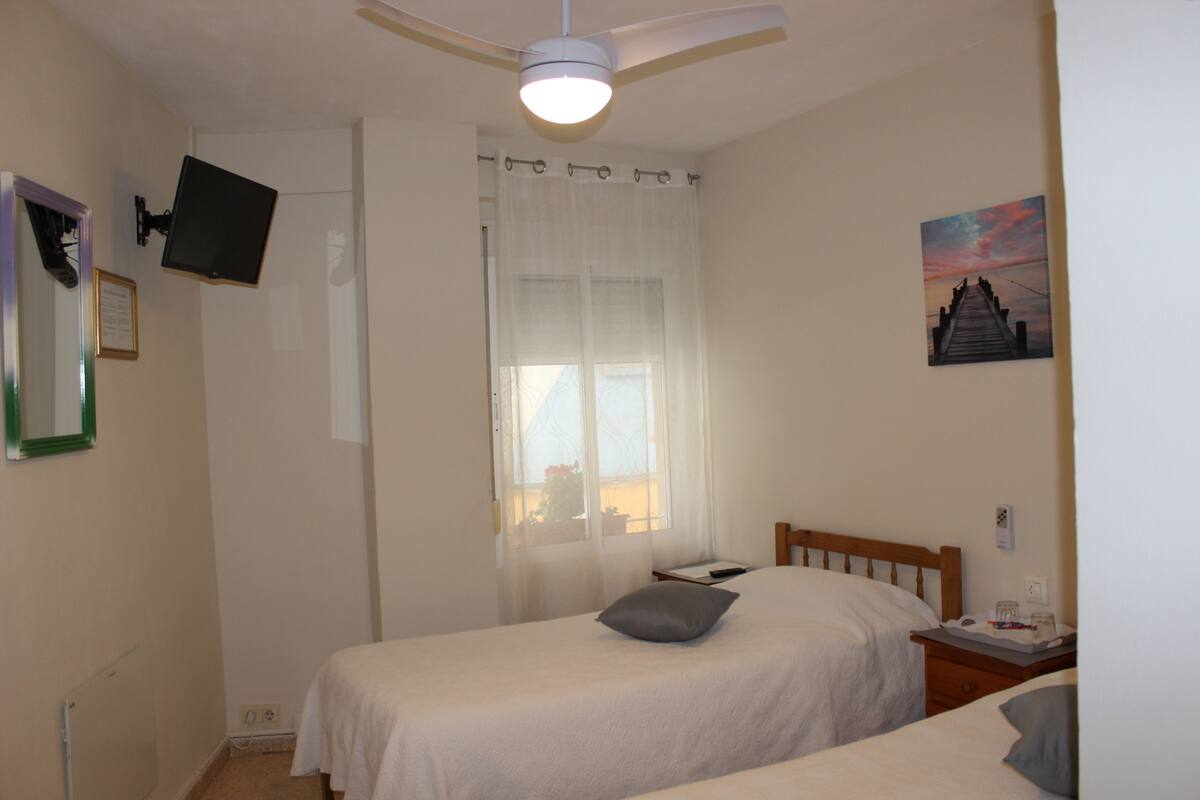 A bedroom with two beds, a ceiling fan, a TV mounted on the wall, a window with sheer curtains, and a painting on the wall.