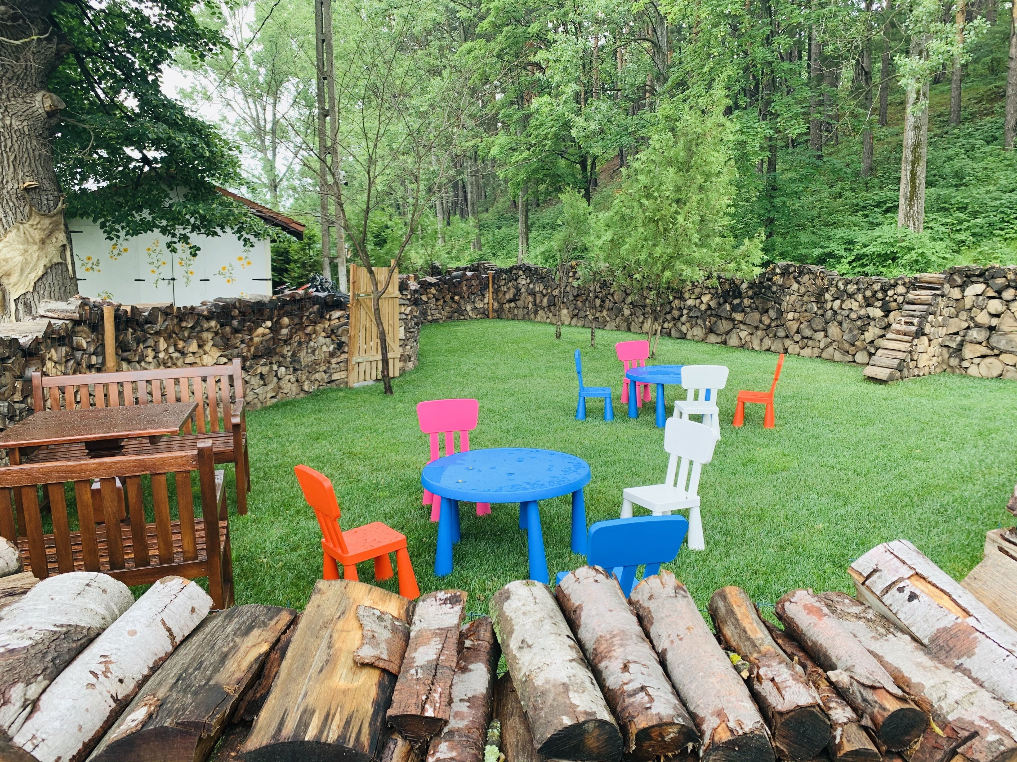 A backyard with a wooden bench, a table, and colorful plastic chairs. There are logs stacked against a stone wall.