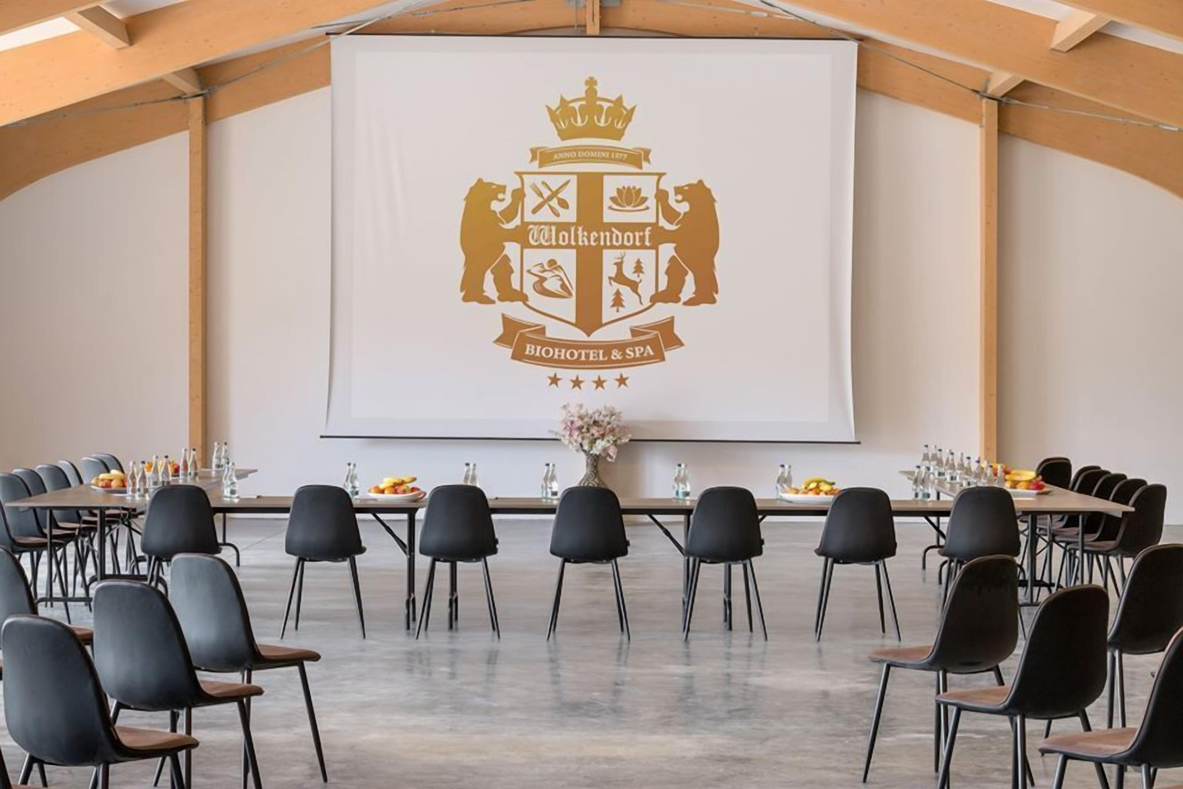 A conference room with a large screen displaying a logo, black chairs arranged around tables, and a vase with flowers on the table.