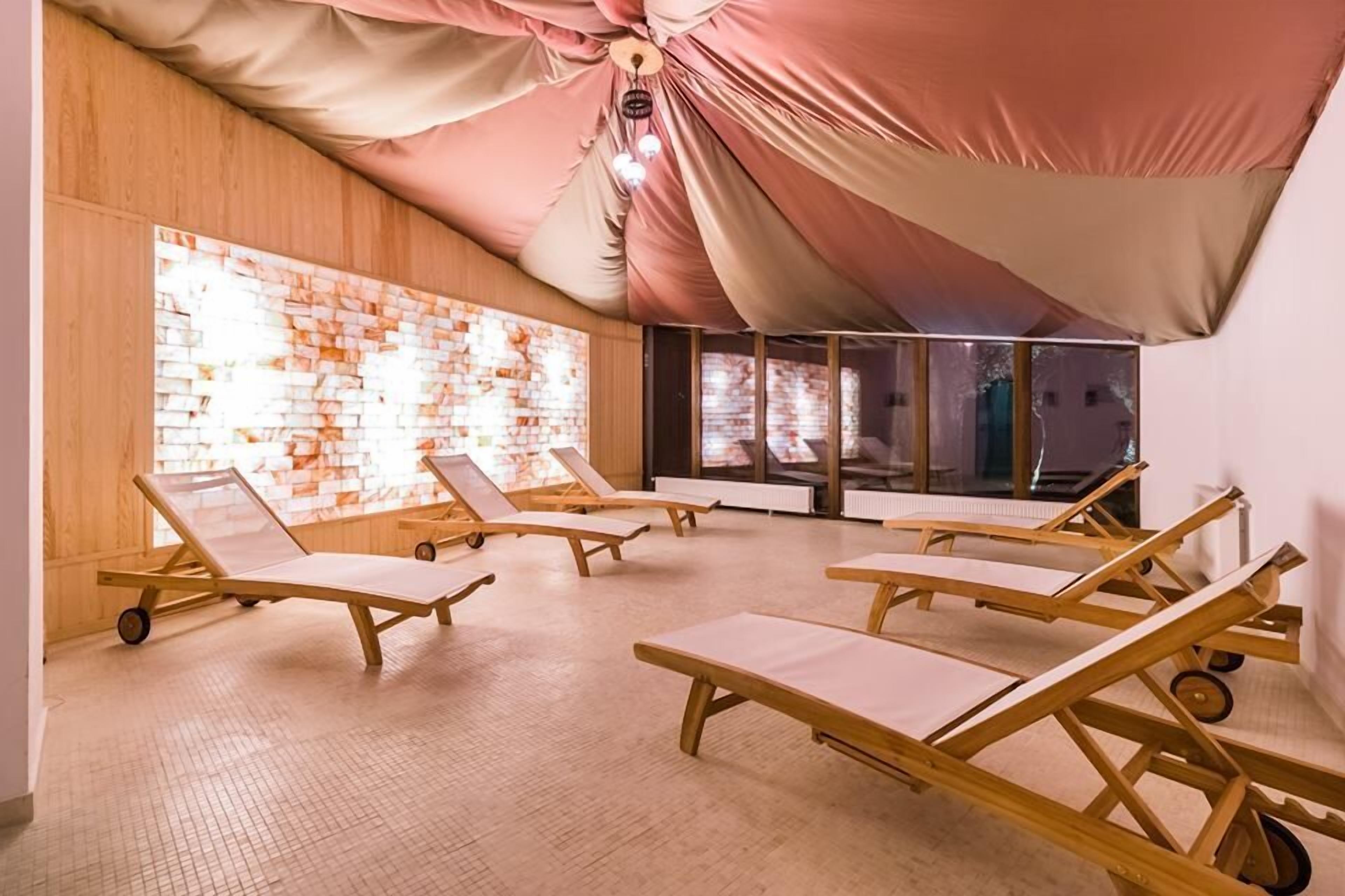 A room with wooden lounge chairs arranged in a semi-circle, a brick wall with a patterned light, and a large window with a view.