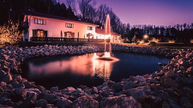 A house with a fountain in a circular stone basin, surrounded by a stone wall, with a forest in the background. - Wolkendorf Bio Hotel & Spa (Vulcan)