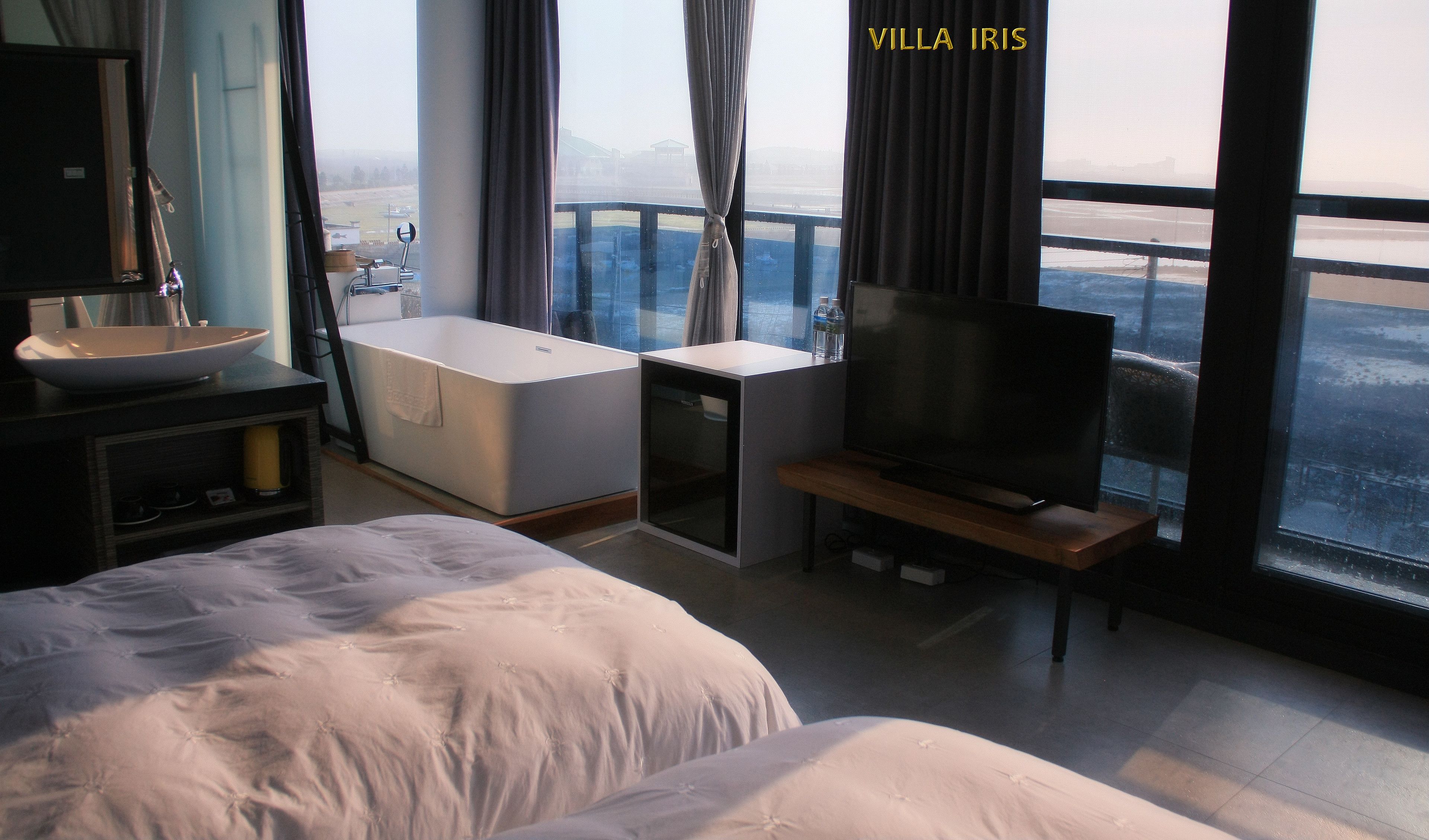 A hotel room with a large bed, a TV on a stand, a small bathroom with a bathtub, and a view of the city through large windows.