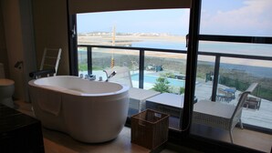A modern bathroom with a large bathtub, a toilet, and a view of a pool and beach through large windows. - Villa Iris (Baisha)