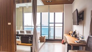 A modern room with a large sliding glass door leading to a balcony with a view of the sea. There is a wooden desk with a chair, a television mounted on the wall, and a small table with a lamp. - Villa Iris (Baisha)