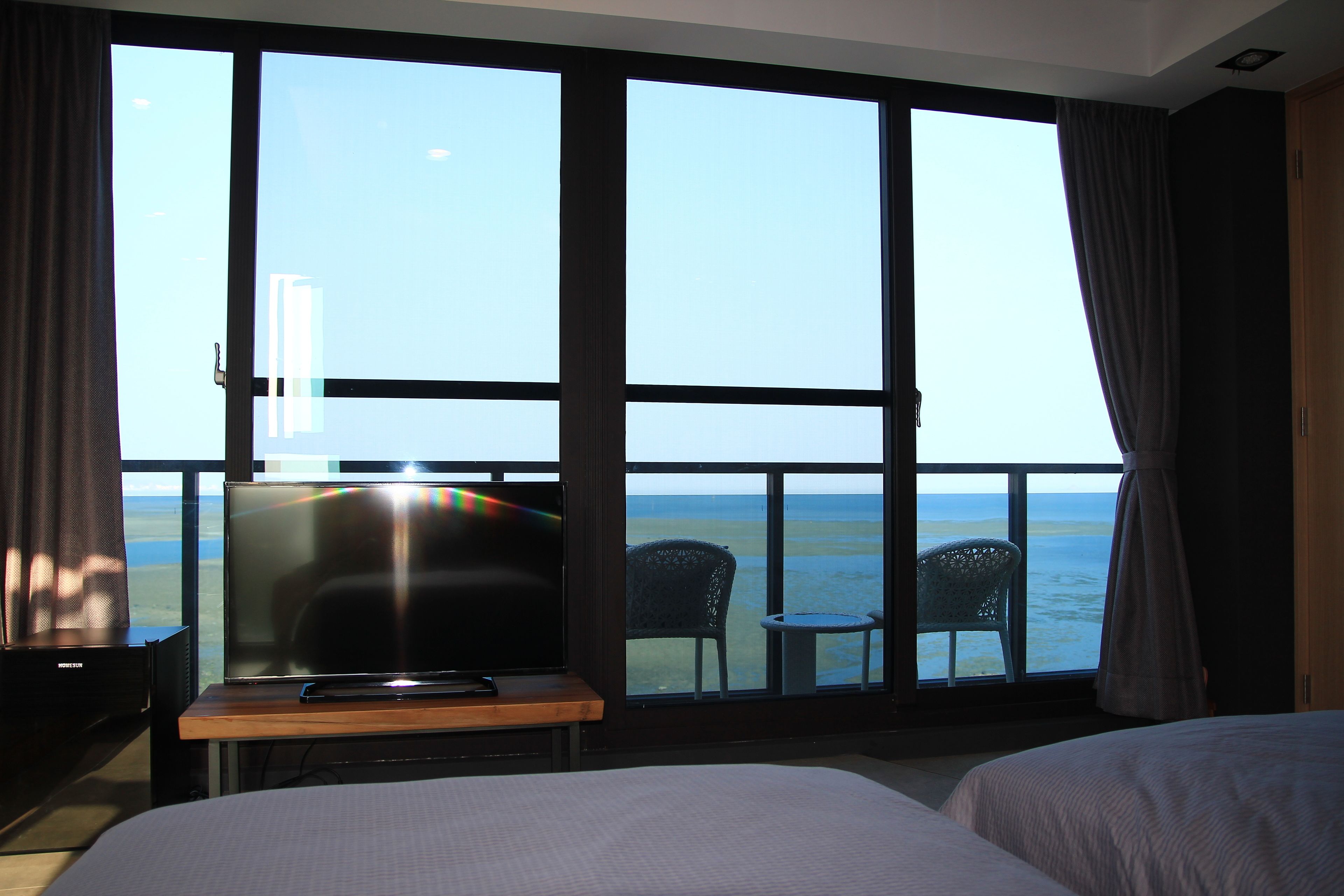 A hotel room with a large window overlooking the sea, a bed, a desk with a TV, and two chairs.