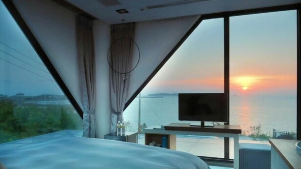A modern bedroom with a large bed, a television, and a view of the sunset over the water. - Villa Iris (Baisha)