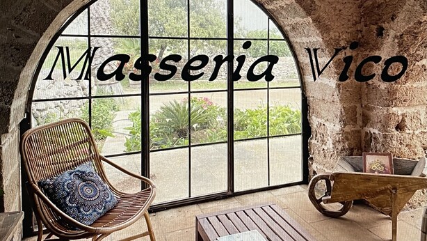 A room with a large arched window, a wicker chair with a patterned cushion, and a wooden cart. - Masseria Vico (Nardò)