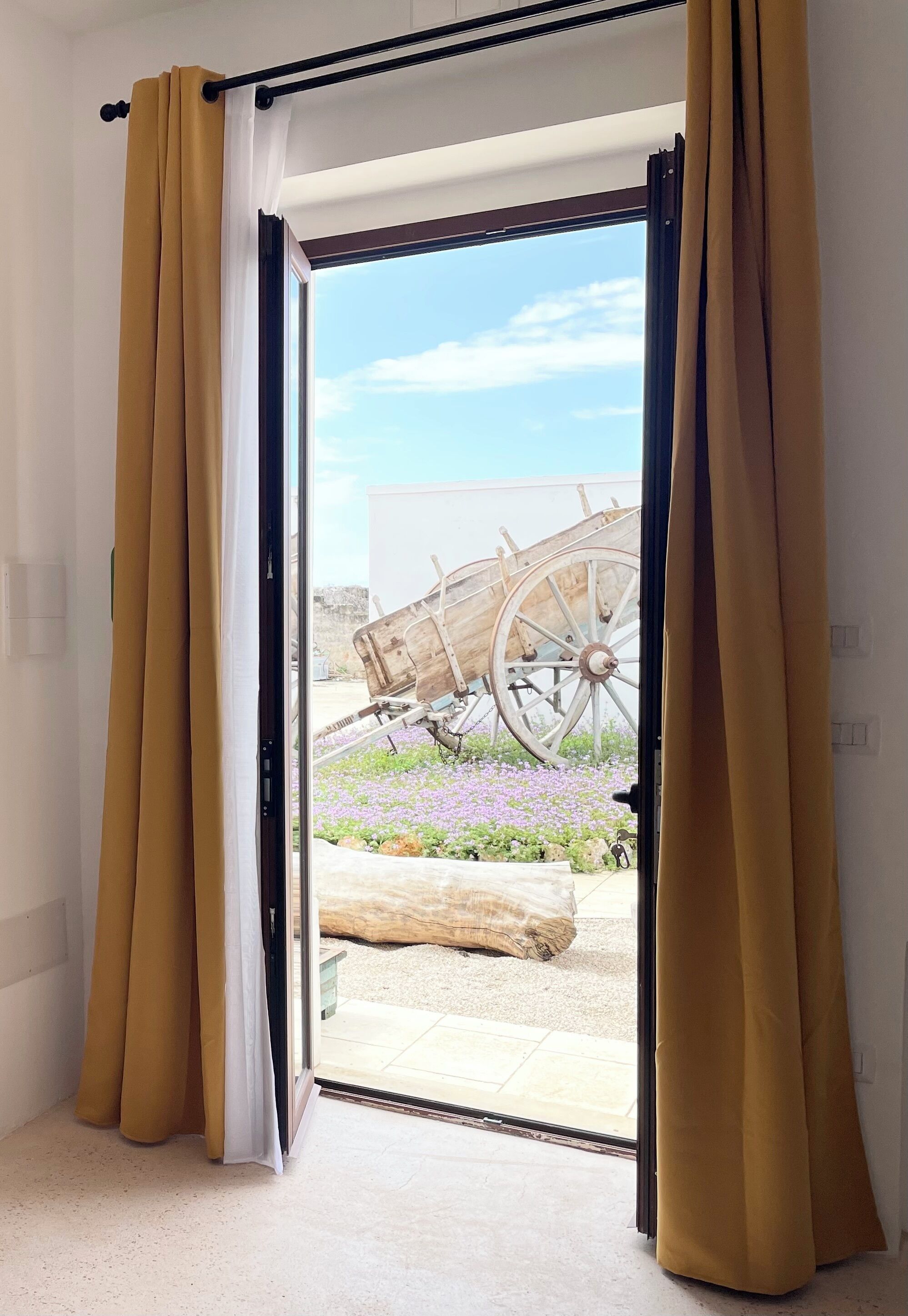 A room with a sliding glass door leading to an outdoor area featuring a wooden cart and purple flowers.