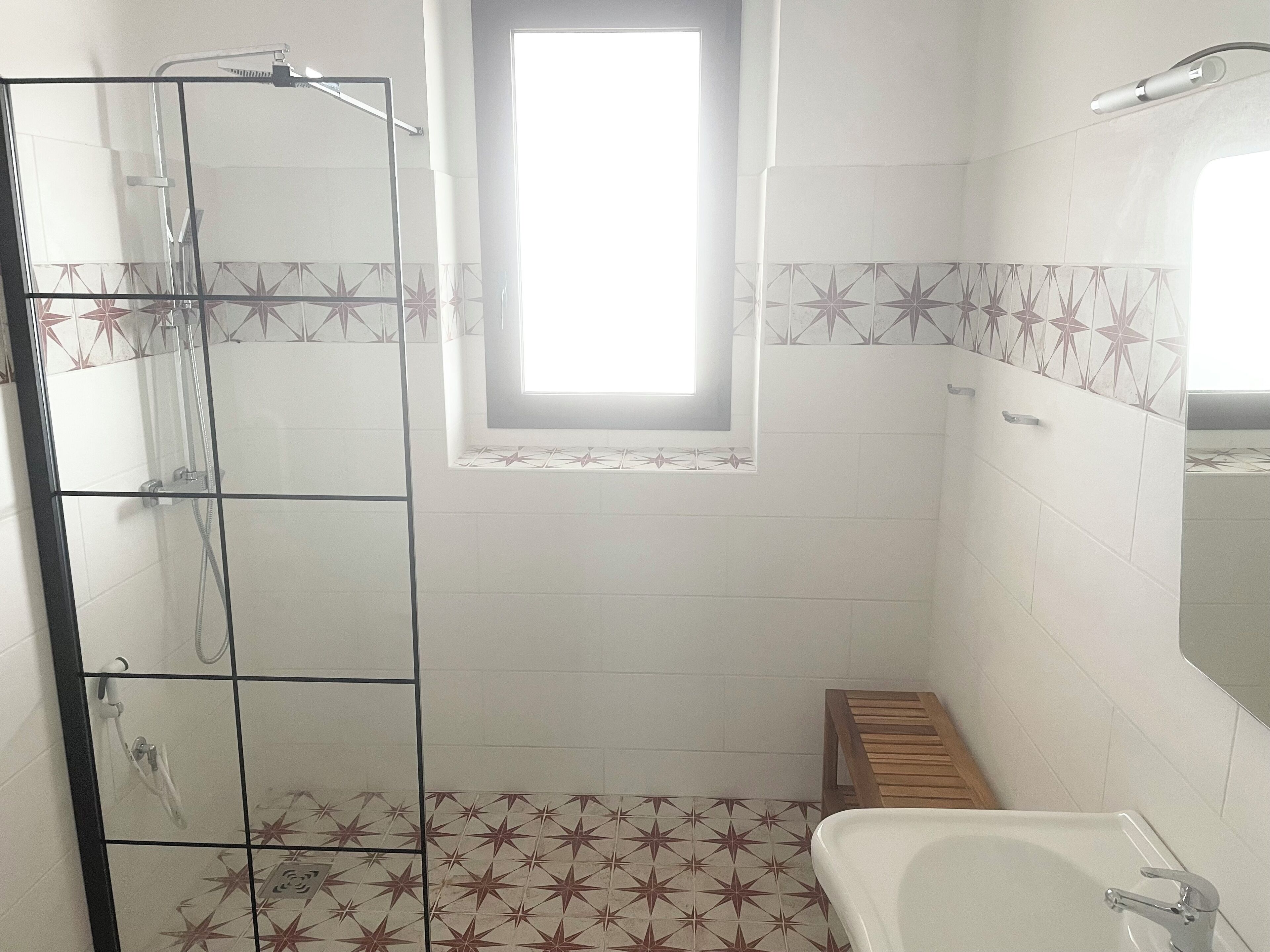 A bathroom with a glass shower enclosure, a small wooden stool, a white bathtub, and a window providing natural light.