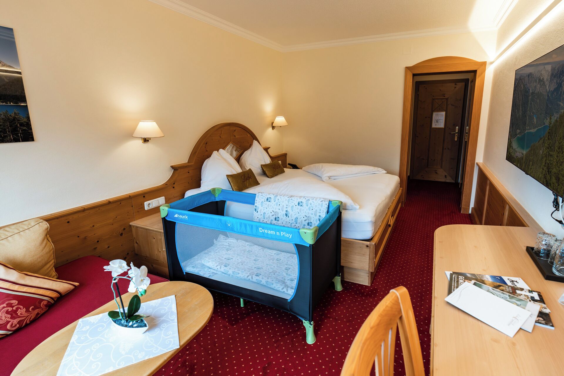 Habitación de hotel con cama, una mesita pequeña con una silla, un escritorio con silla, una lámpara de pared y una puerta que da a otra habitación.
