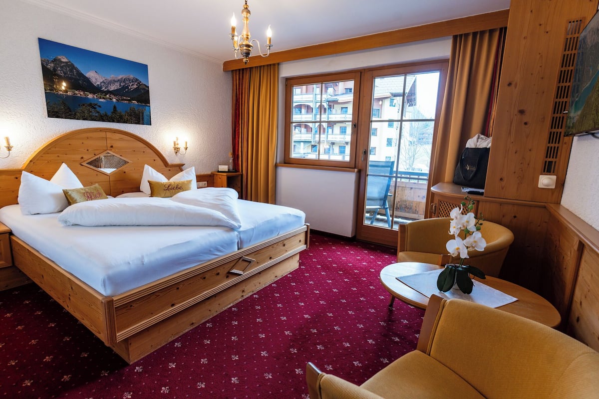 A hotel room with a large bed, a wooden headboard, a small table, a chair, a view of buildings through a window, and a painting on the wall.