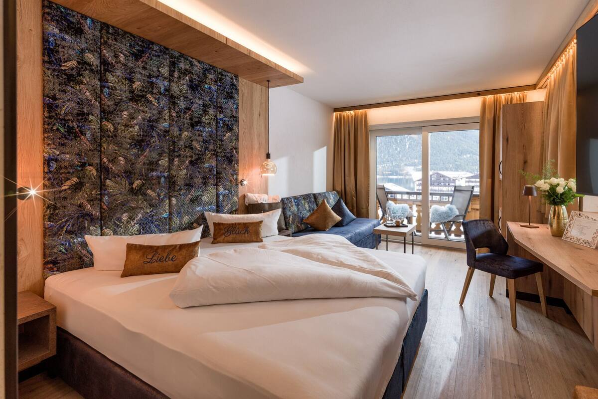 A modern hotel room with a large bed, a blue sofa, a wooden desk, and a view of the mountains through the window.