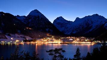 A lakeside town at night with illuminated buildings and snow-covered mountains in the background.