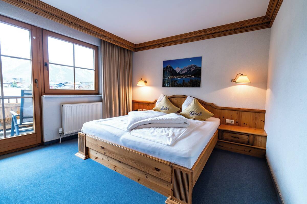 A hotel room with a wooden bed, two bedside lamps, a window with curtains, and a painting on the wall.