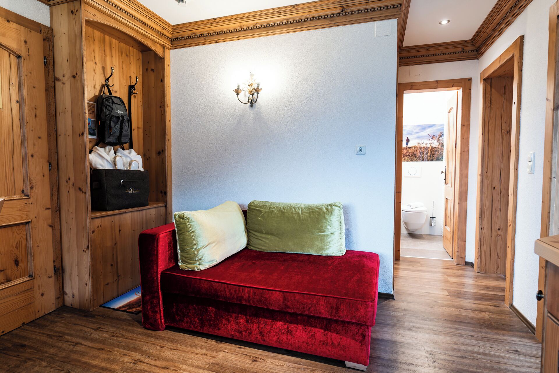 A red velvet sofa with two cushions, a wooden wardrobe, and a wall-mounted light fixture in a room with wooden floors and a view of the outdoors.