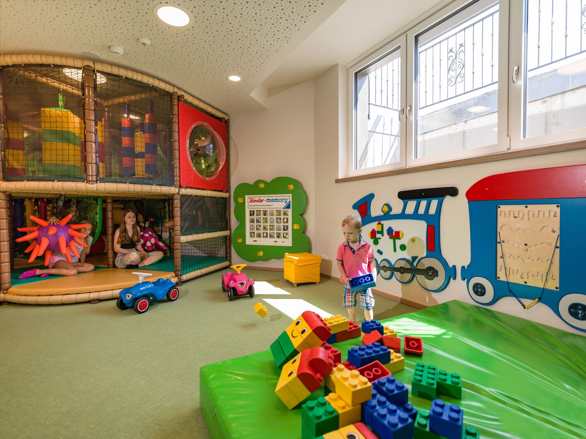 Zona de juegos infantiles con una estructura para escalar, un muro con temática de trenes y una variedad de juguetes.