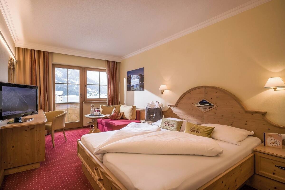 A hotel room with a large bed, a desk with a TV, a small table, and a view of mountains through the window.