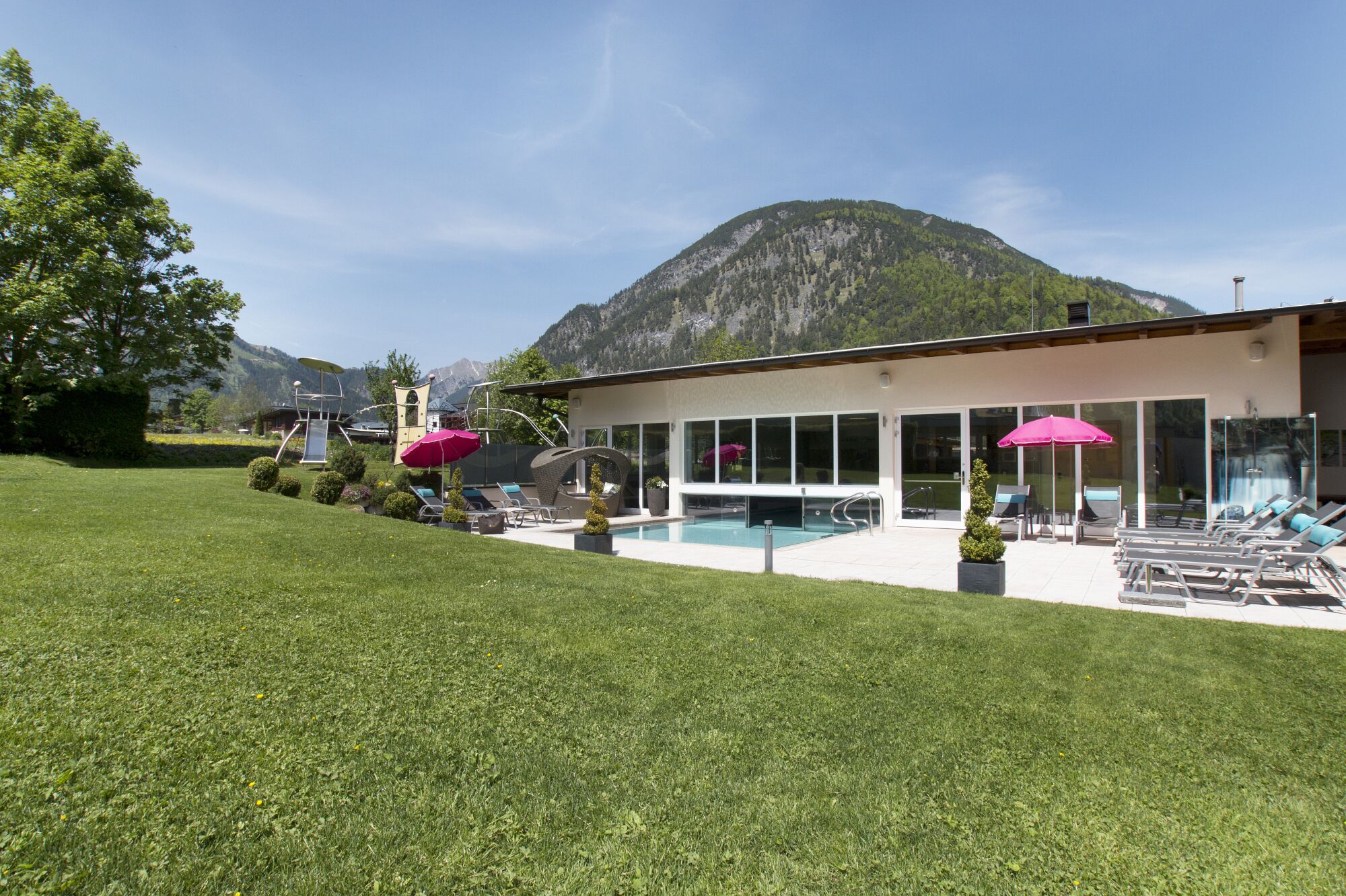 A modern building with a swimming pool, sun loungers, and a mountain backdrop.